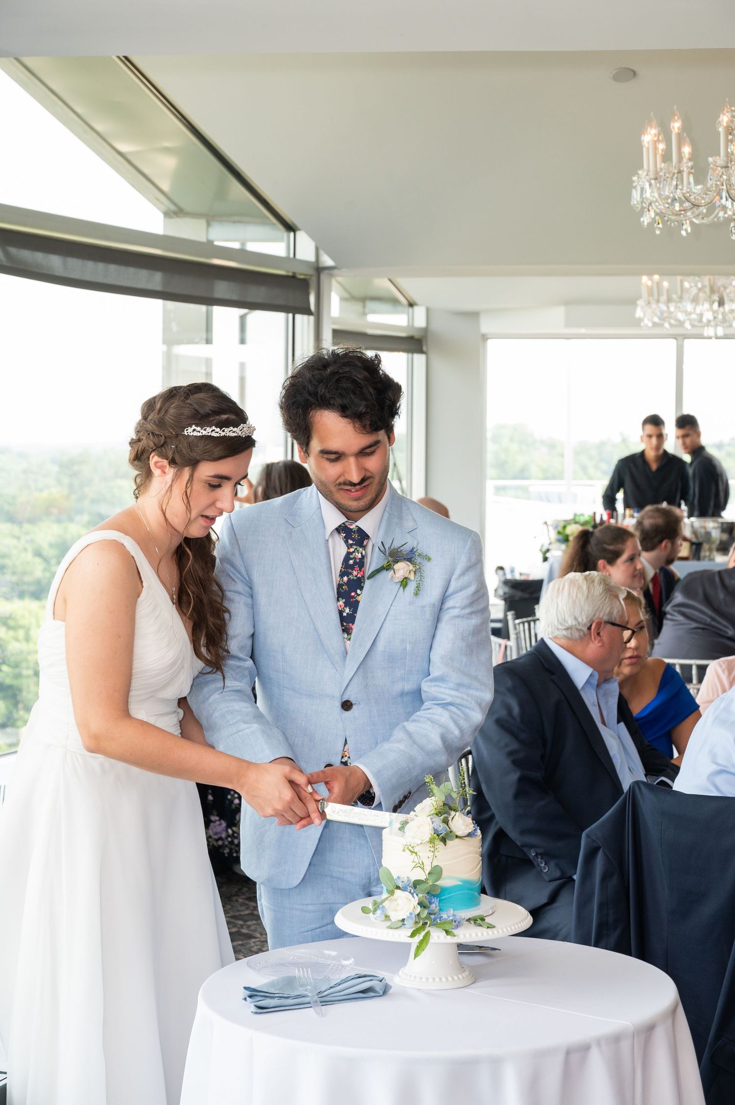 Bride and groom cutting their small wedding cake at a reception venue with guests seated nearby.