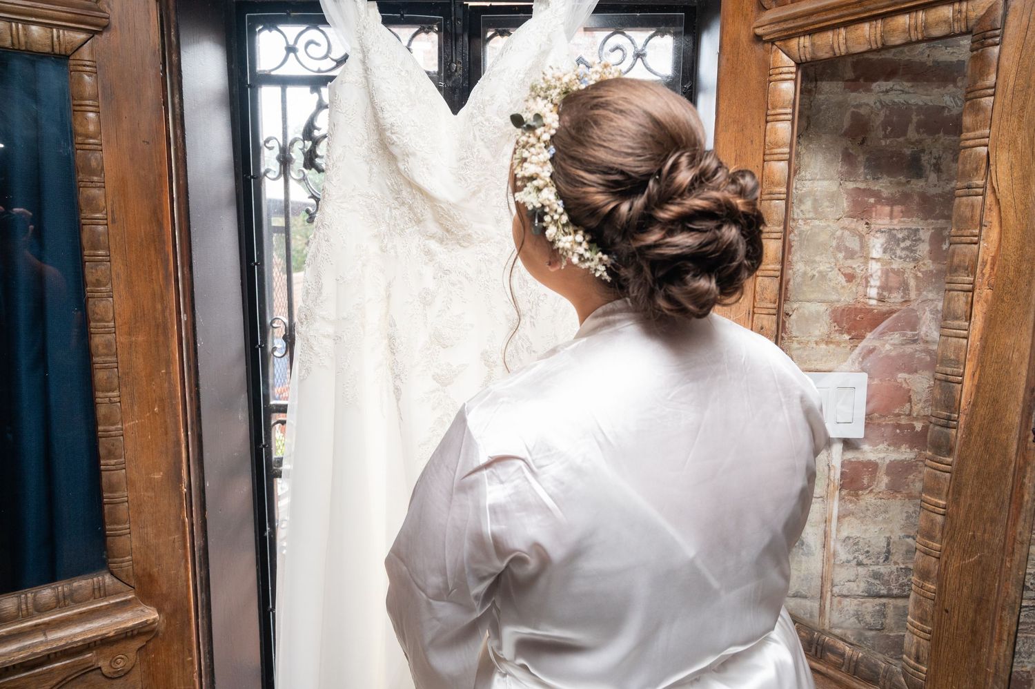 Bride in white robe with elegant updo viewing her lace wedding dress on door.