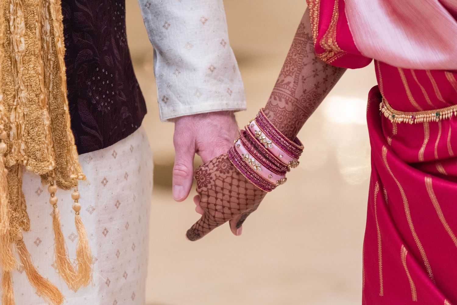 Indian bride and groom holding hands, bride wearing henna, bangles, and red bridal attire during traditional wedding ceremony.