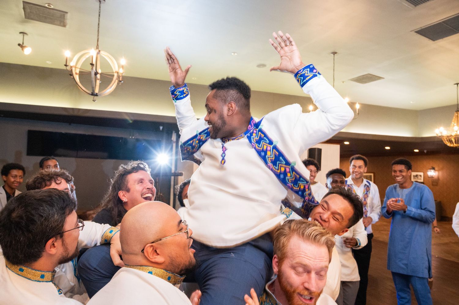 Groom in white and blue traditional attire being lifted by groomsmen during joyful wedding reception celebration.