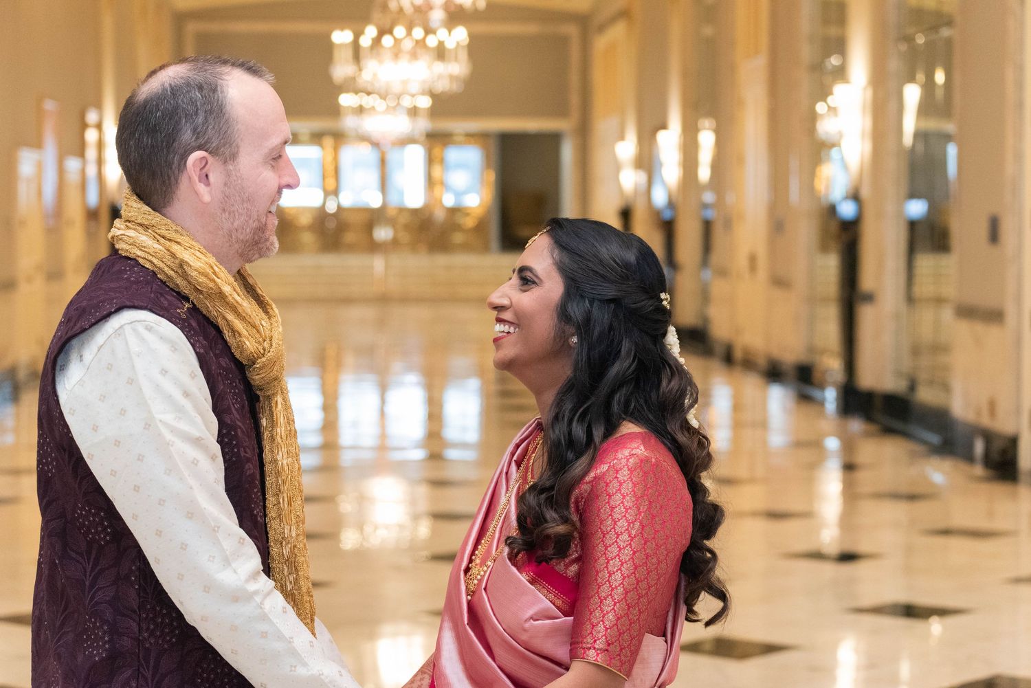Couple in traditional Indian wedding attire smiling at each other in an elegant chandelier-lit hall.