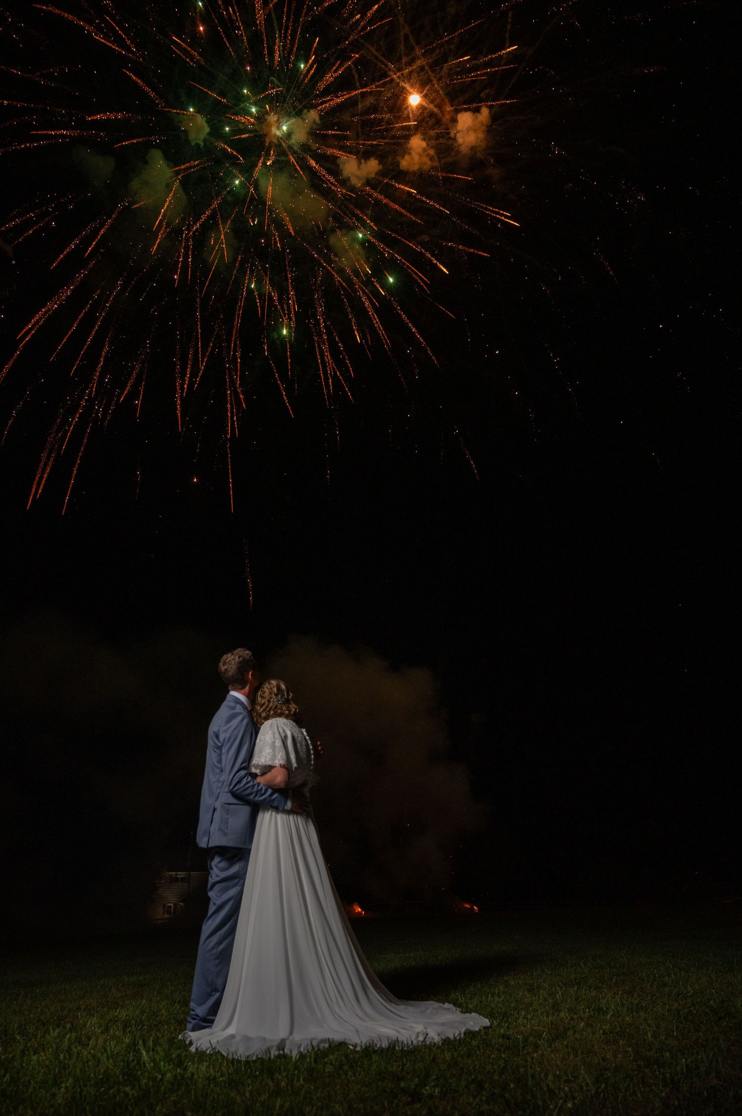 Newlyweds embrace while watching colorful fireworks display light up the night sky at outdoor wedding.