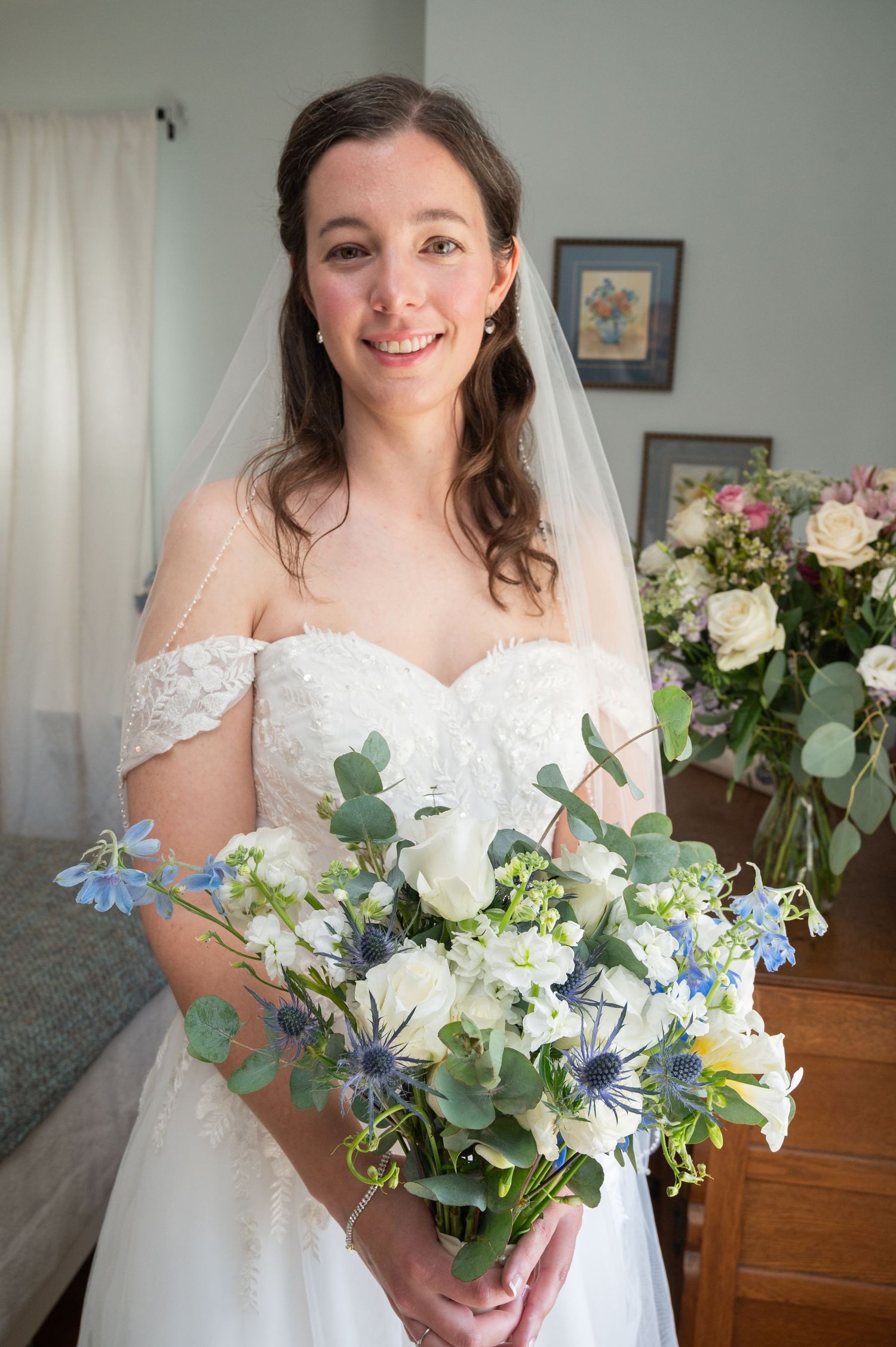 Smiling bride in off-shoulder lace gown holding blue and white floral bouquet with veil.