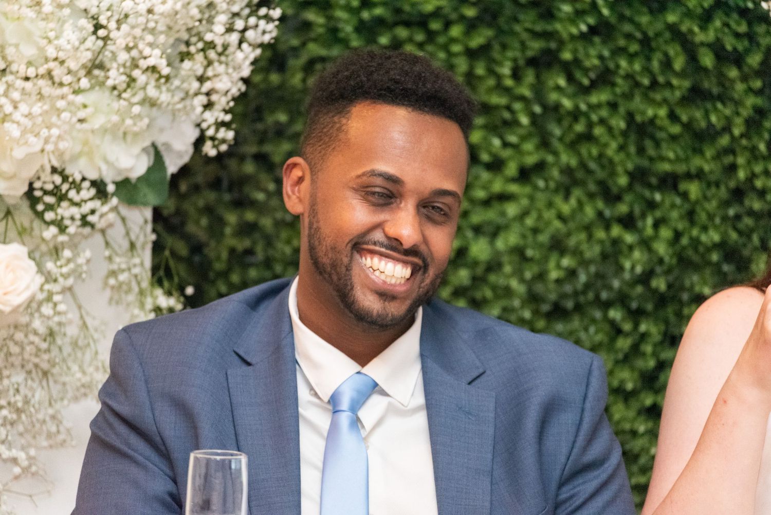 Smiling man in blue suit and tie at a wedding reception with floral backdrop.