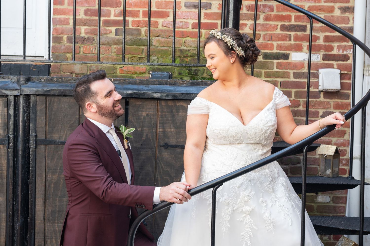 Groom in burgundy suit holds bride's hand on iron staircase outside brick building wedding portrait.