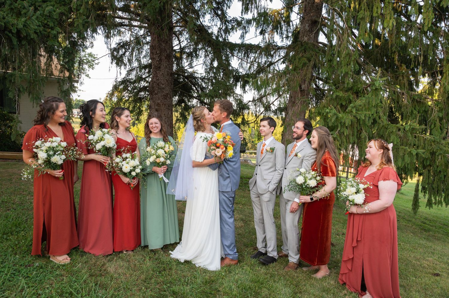 Wedding party in rust and sage dresses surrounds kissing couple under tall pine trees outdoors.