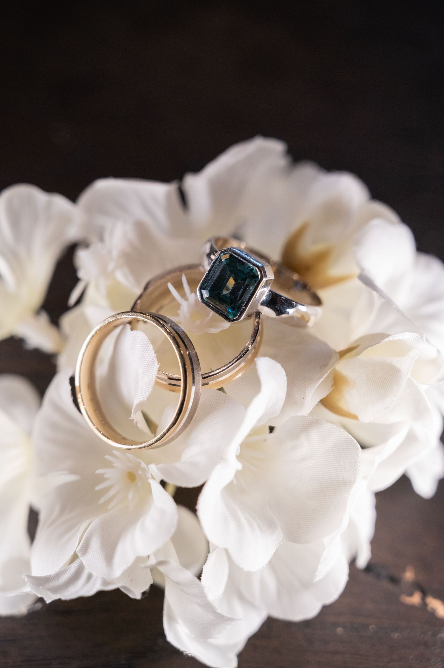 Wedding rings rest on white flowers, featuring a gold band and a silver ring with a dark gemstone.