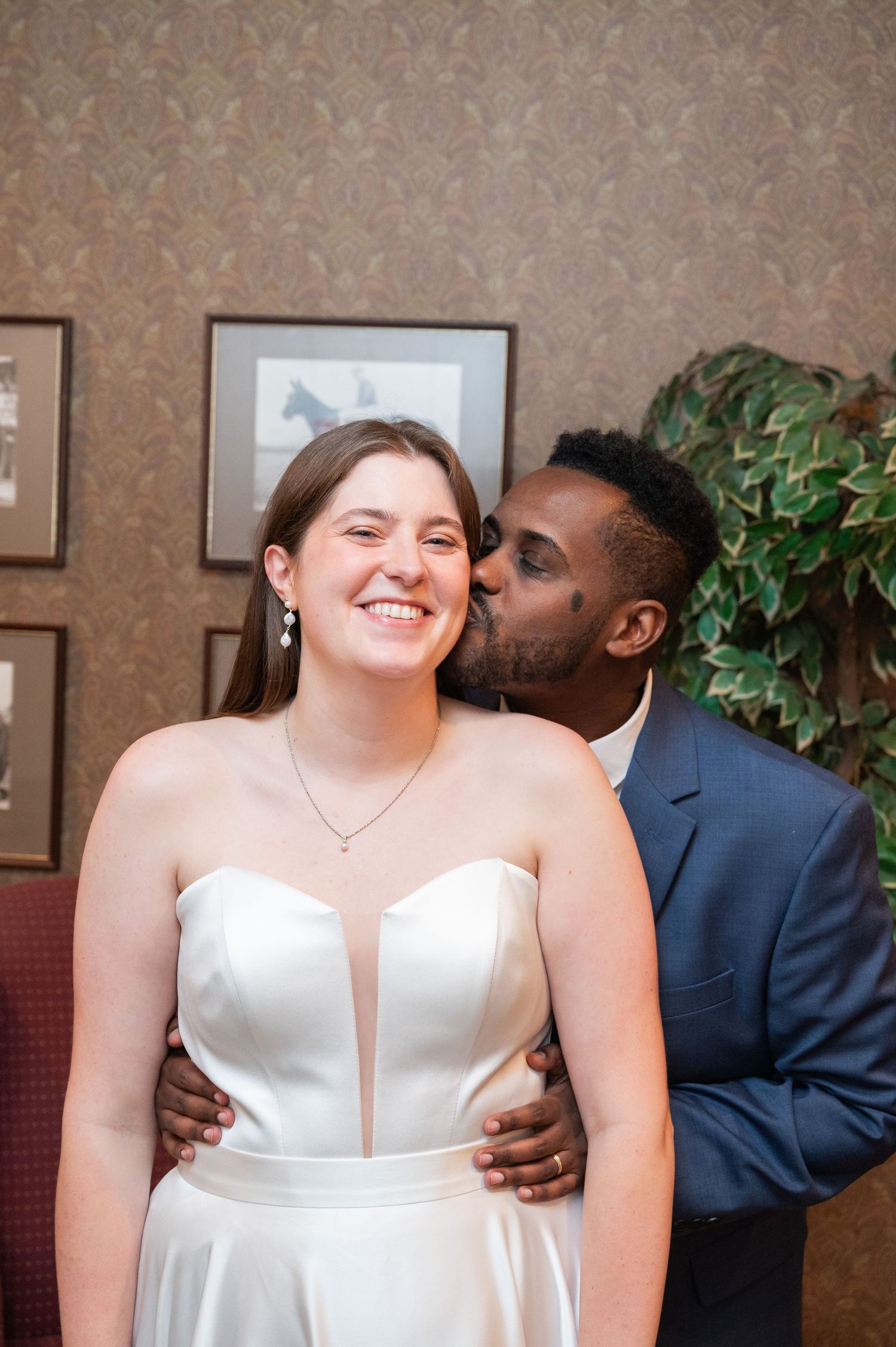 Smiling bride in white strapless gown receives a kiss on the cheek from groom in blue suit.