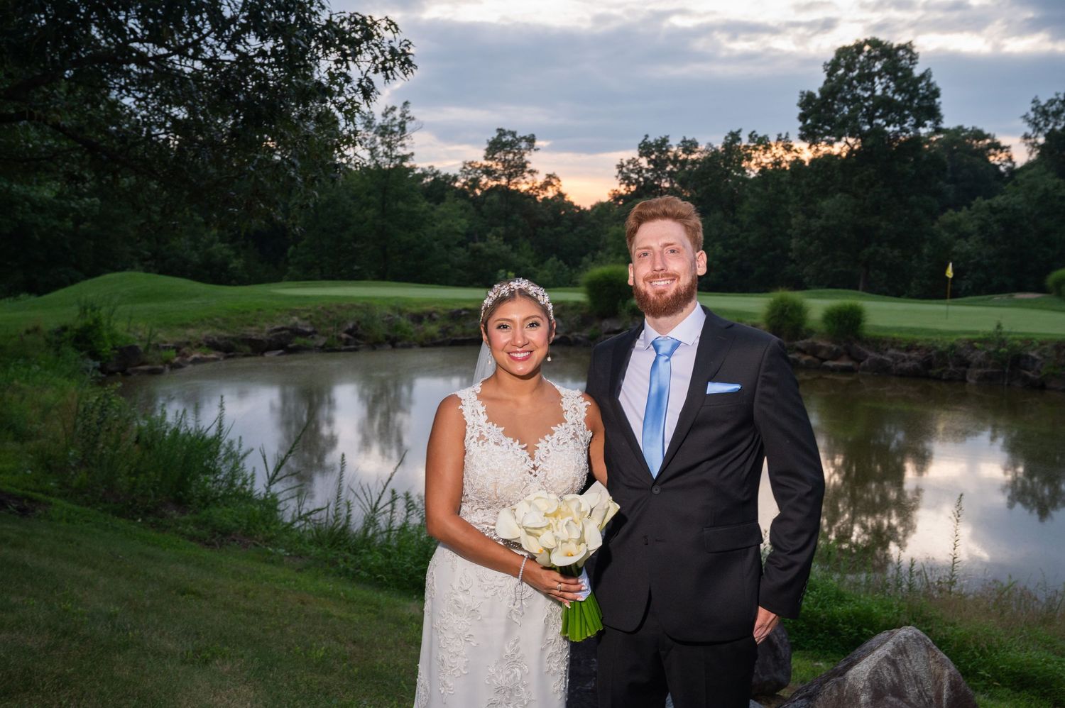 Bride and groom posing together at sunset near a pond on a golf course, bride holding white bouquet.