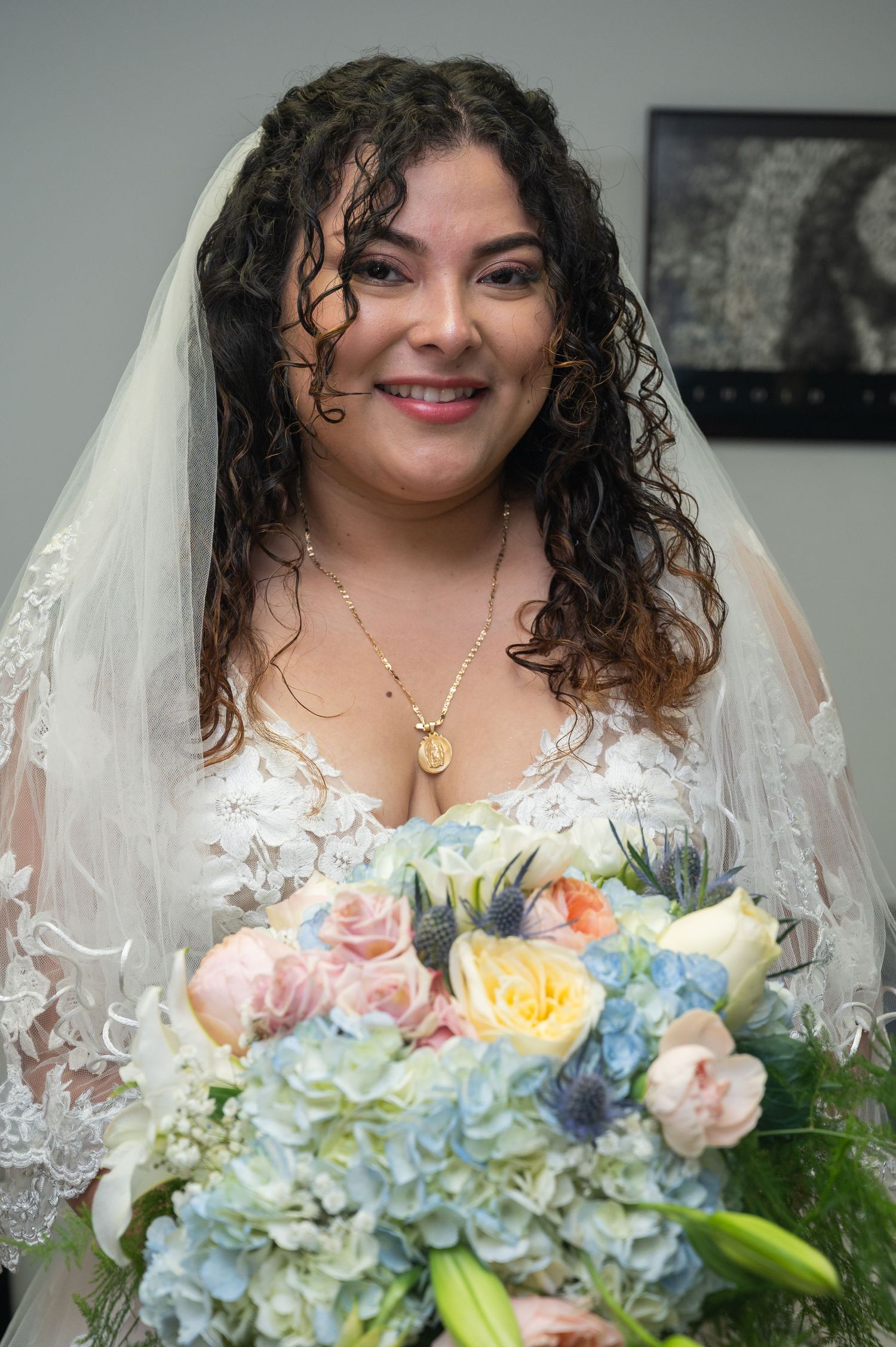 Curly-haired bride in lace gown holds pastel bouquet with blue hydrangeas, wearing gold necklace.