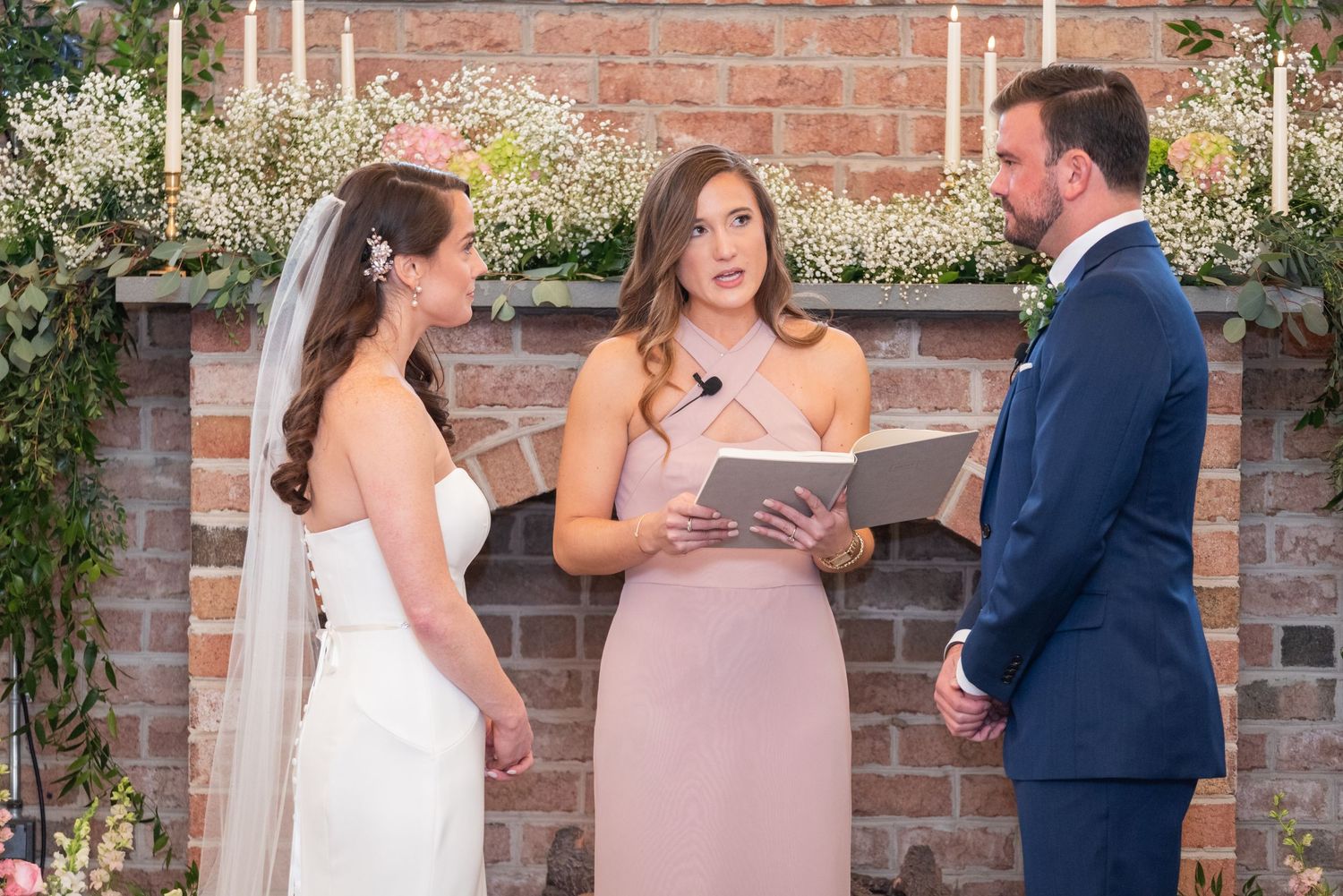 Wedding officiant reads vows as bride and groom face each other at rustic brick fireplace altar.
