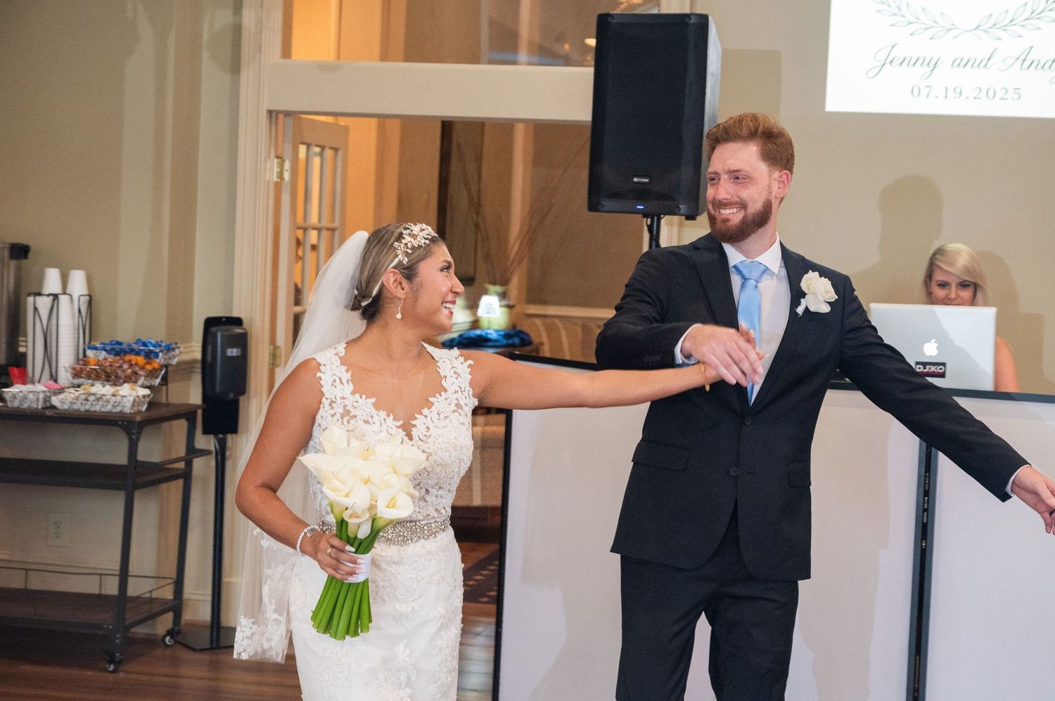 Bride in white lace gown holding bouquet and groom in black suit smiling during wedding reception entrance.