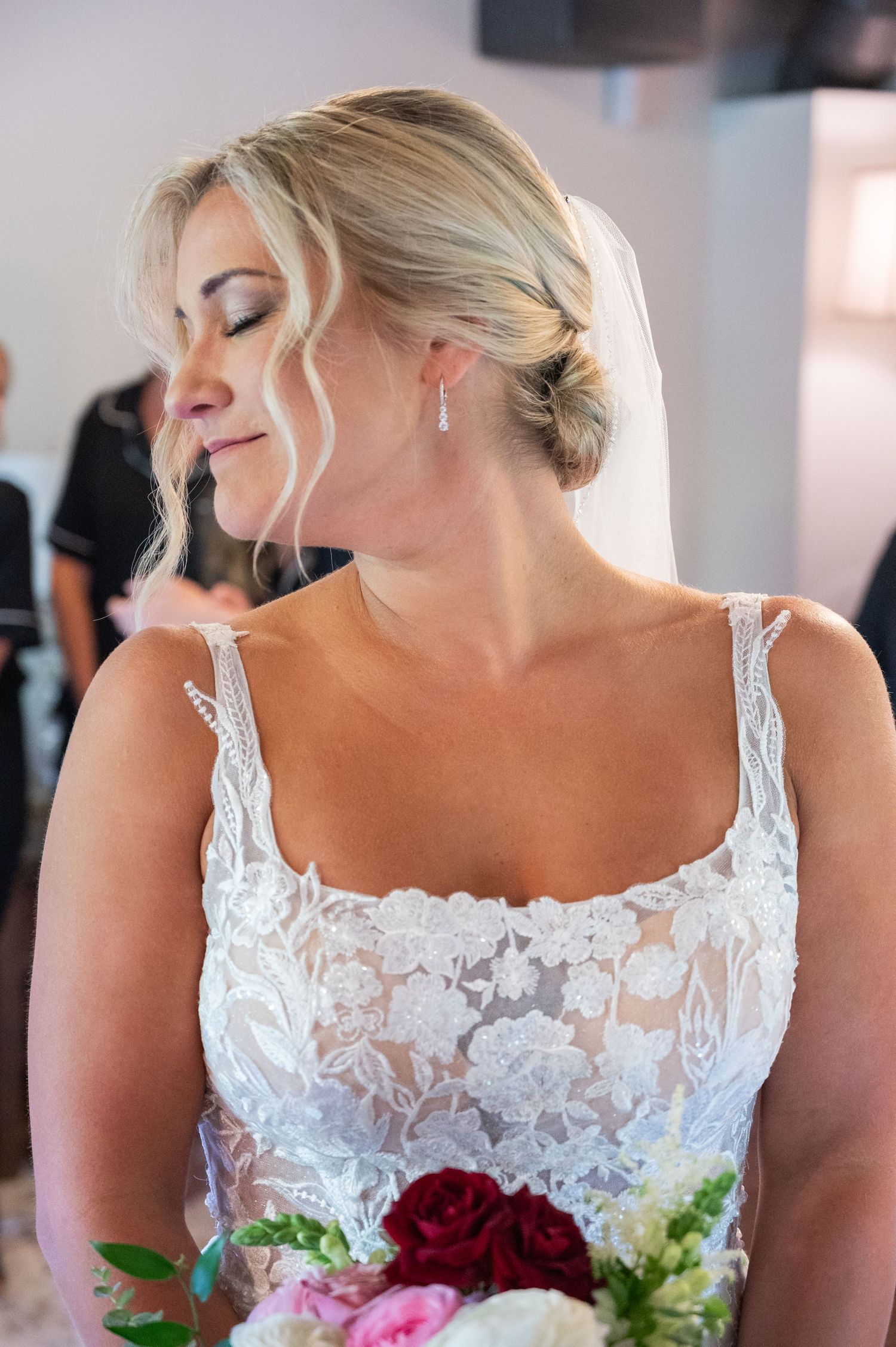 Blonde bride in lace wedding gown holds bouquet with eyes closed, wearing drop earrings and veil.