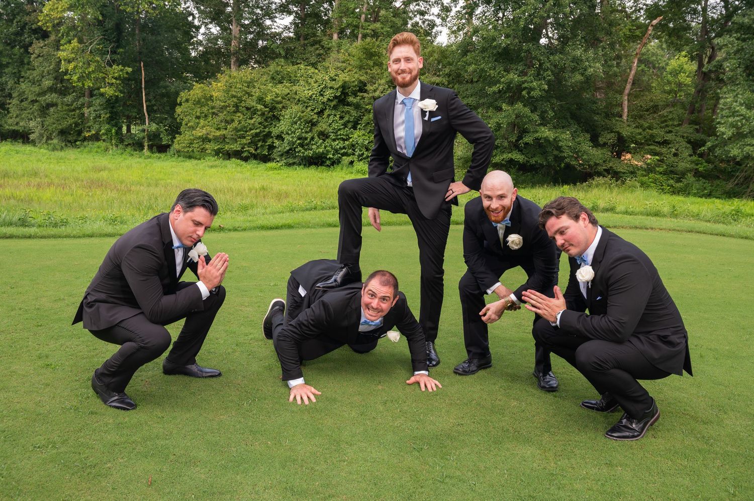 Five groomsmen in black suits posing playfully on a golf course green with lush trees in the background.