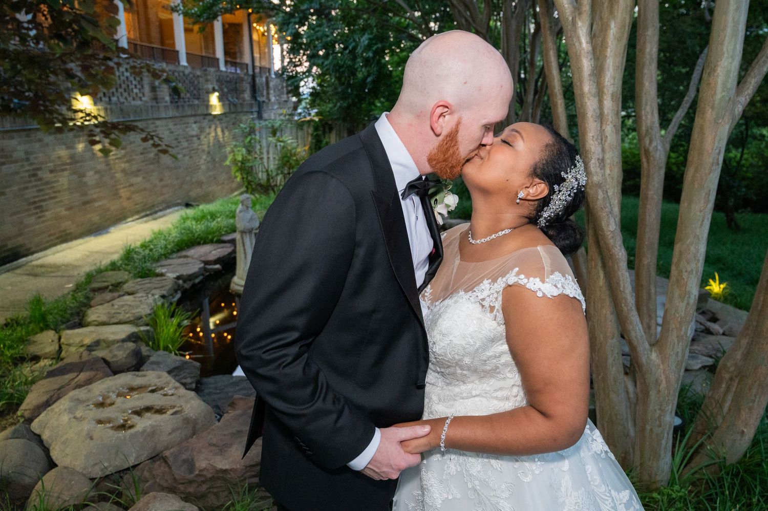 Interracial couple sharing a romantic kiss in wedding attire outdoors near a stone garden backdrop.