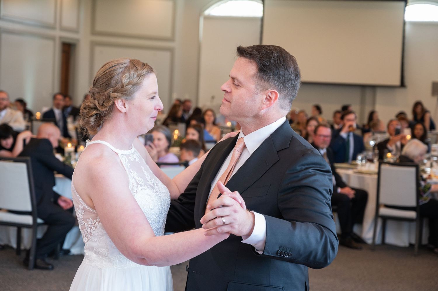 Bride and groom sharing their first dance at a wedding reception with guests watching nearby.