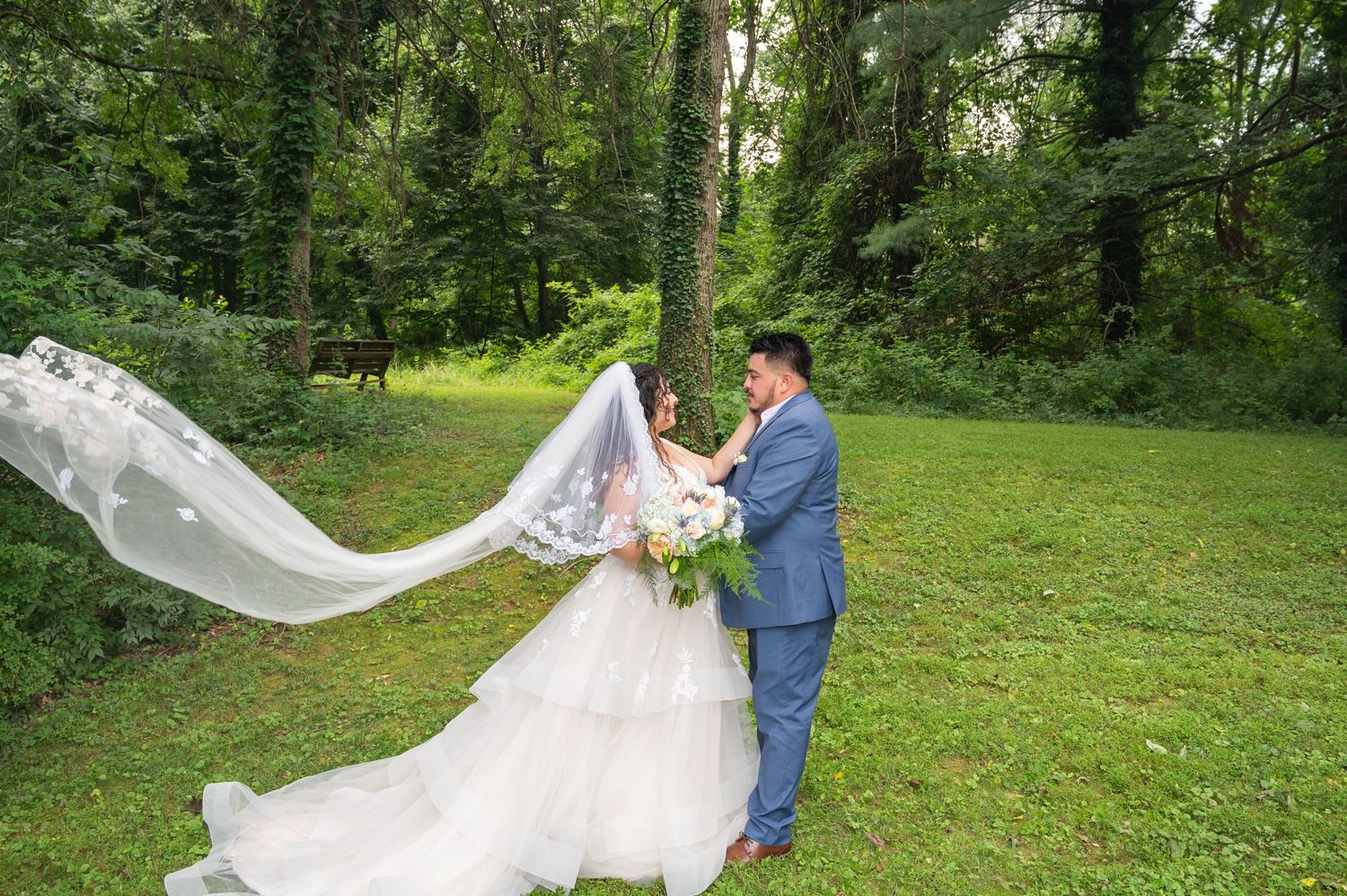 Bride in flowing gown with dramatic veil dancing with groom in gray suit in lush forest clearing.