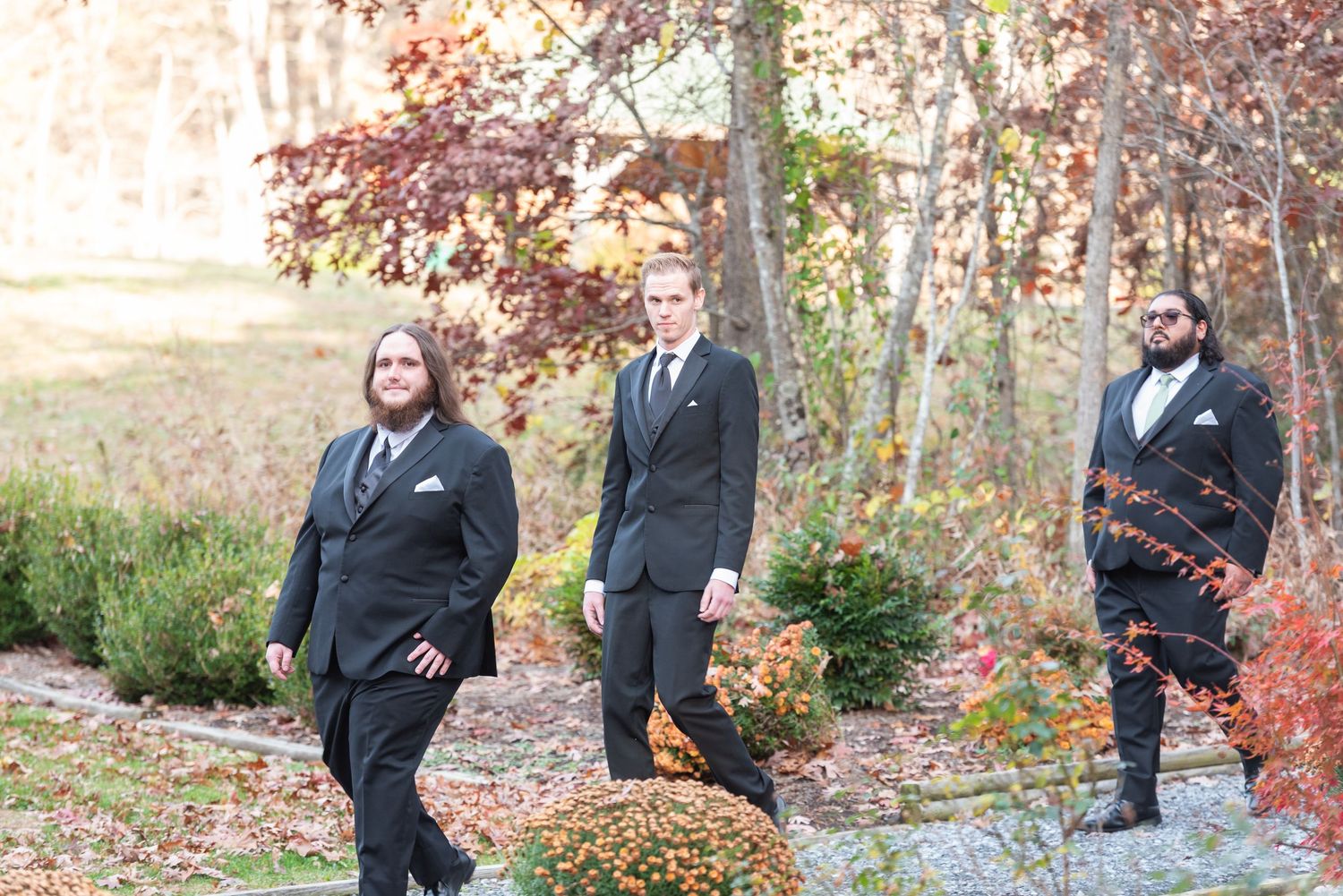 Three groomsmen in black suits posing outdoors among autumn foliage and stone pathway.