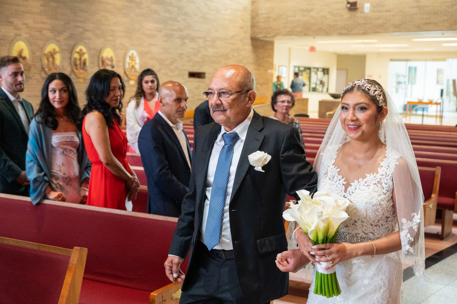 Bride in white lace gown and veil walks down church aisle with older man, carrying white calla lily bouquet.