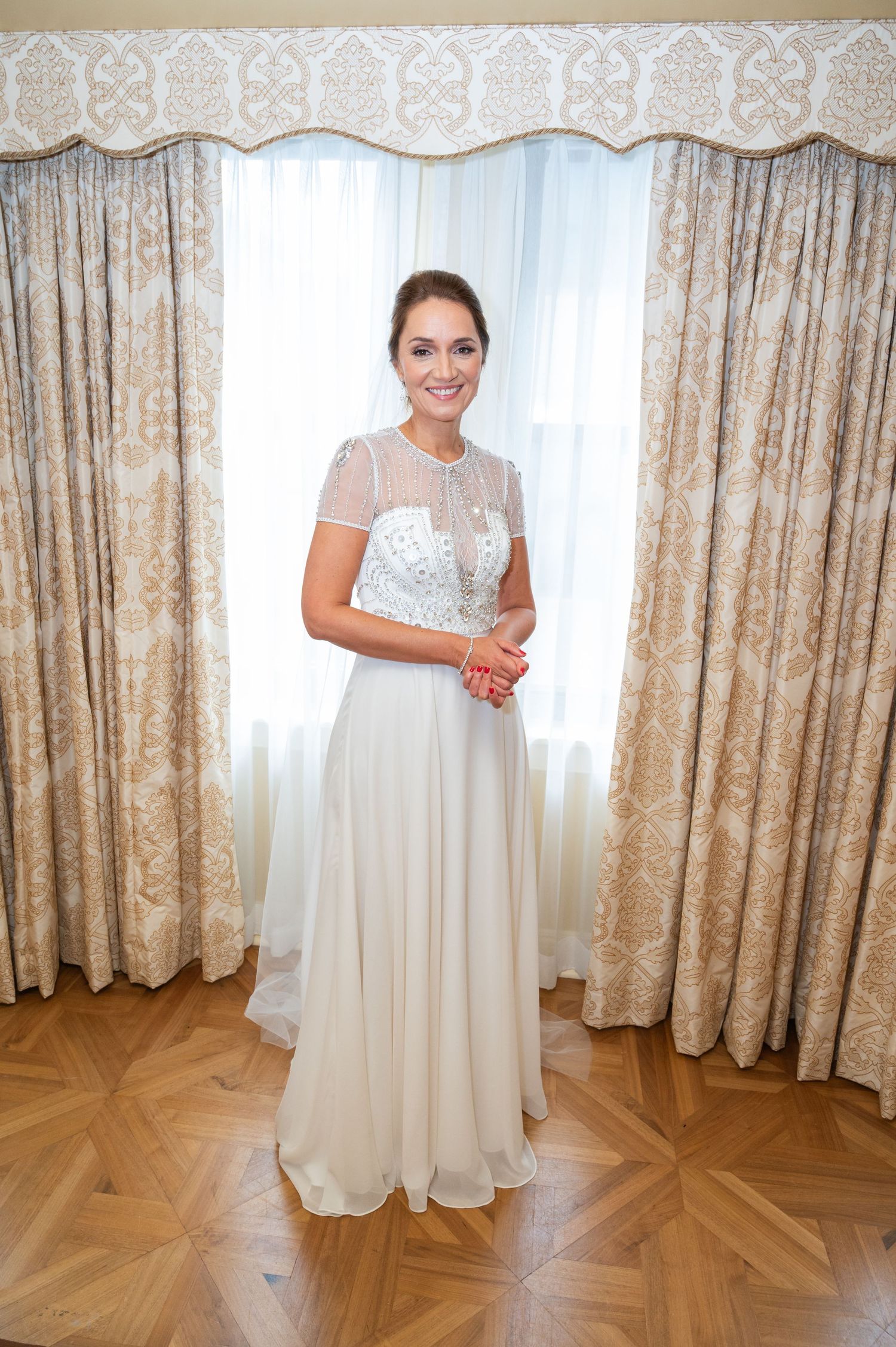 Radiant bride in beaded illusion bodice wedding gown standing before ornate curtains.