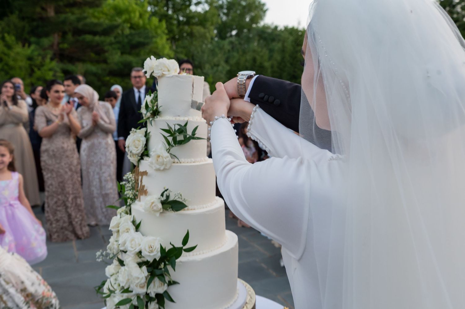 Bride and groom cutting a tall white five-tier wedding cake decorated with white flowers outdoors.