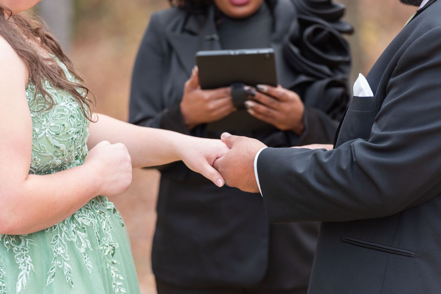Wedding officiant holding tablet while couple exchanges vows, bride in green gown holding grooms hands.