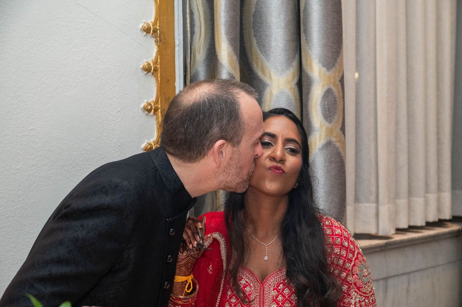 Man kissing woman on cheek at a festive celebration, woman wearing red embroidered outfit.