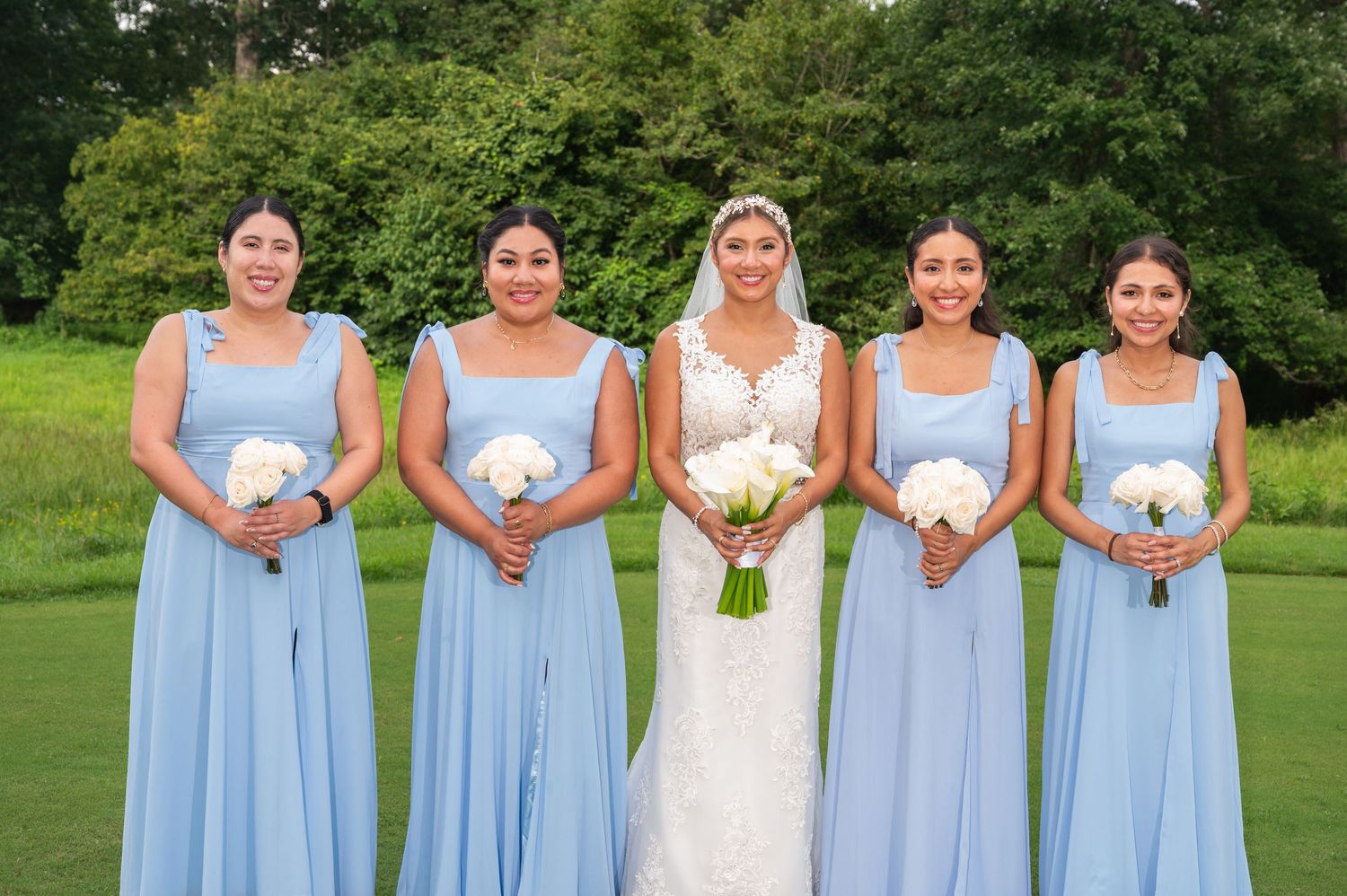 Bride in white lace gown with four bridesmaids in light blue dresses holding white bouquets outdoors.