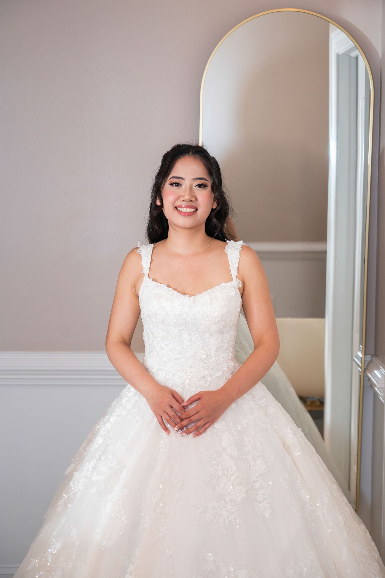 Smiling bride in a white lace ball gown wedding dress standing in front of a mirror in a bridal boutique.