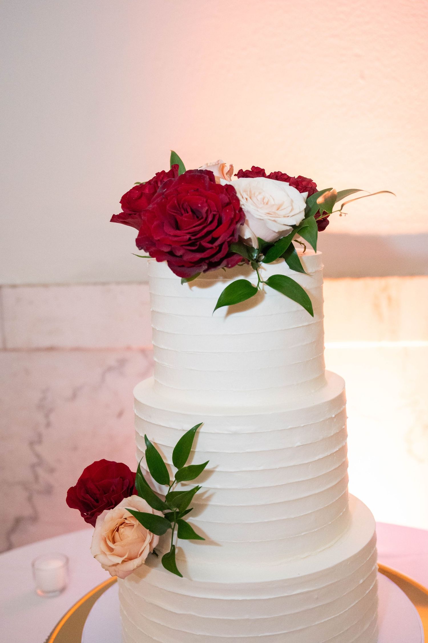 Elegant three-tier white wedding cake adorned with red and blush roses and green leaves.