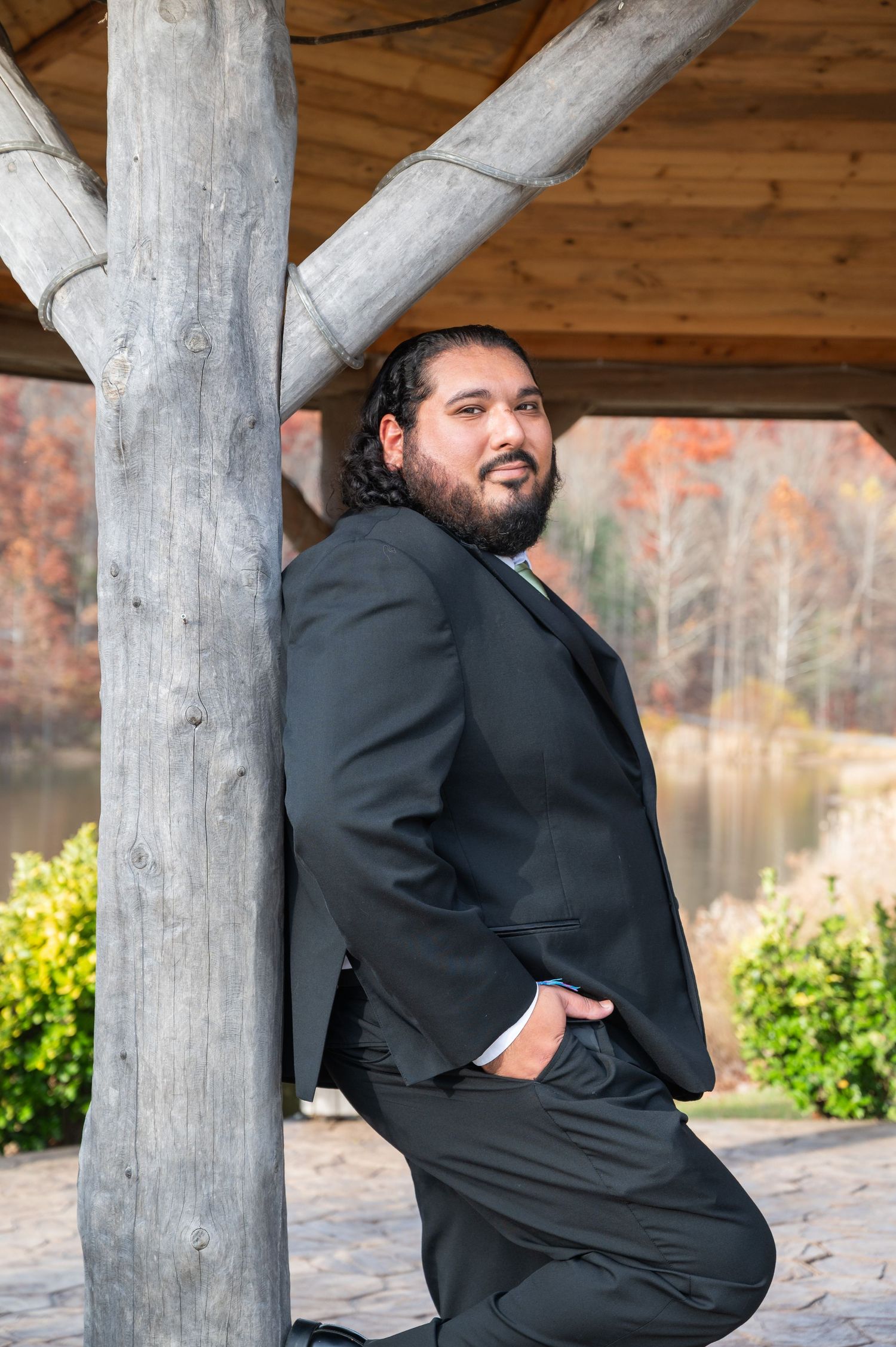 Groom in black suit leaning against rustic wooden pavilion post with autumn foliage and pond in background.