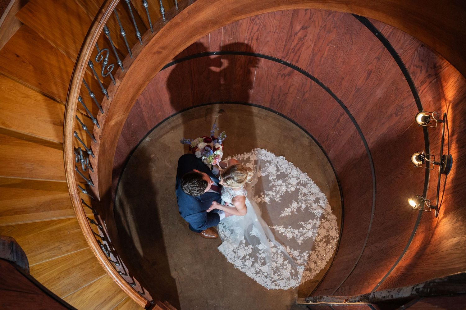 Top-down view of wedding couple sitting beneath a stunning circular wooden spiral staircase.