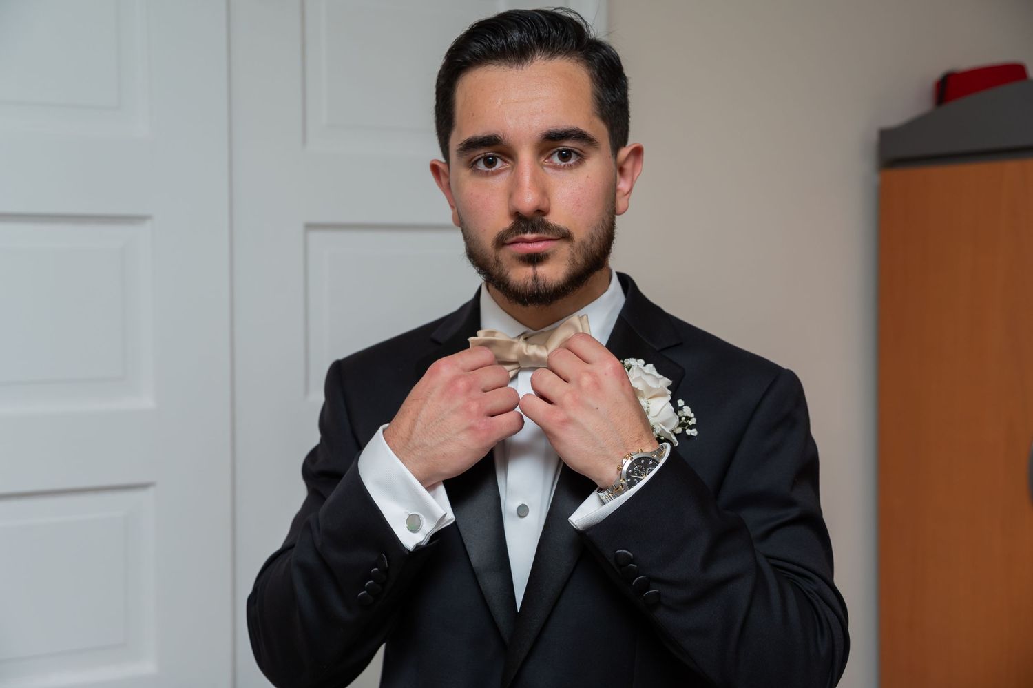 A well-dressed groom in a black suit adjusts his bow tie while wearing a boutonniere and silver watch.