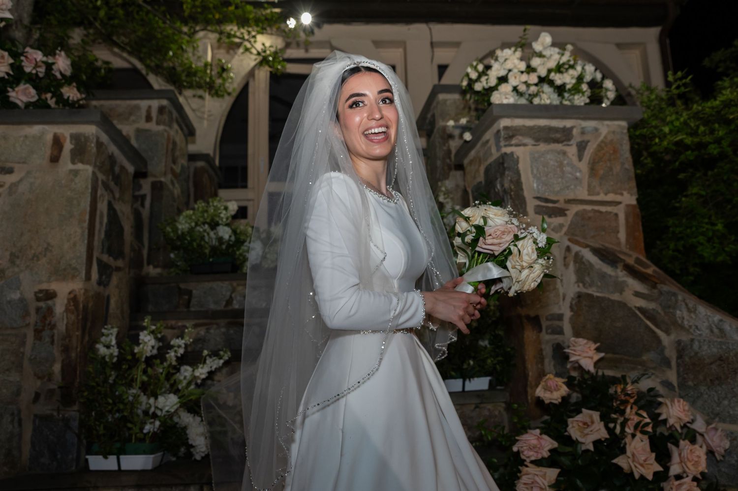 Smiling bride in white gown and veil holding bouquet at floral-decorated stone church entrance.