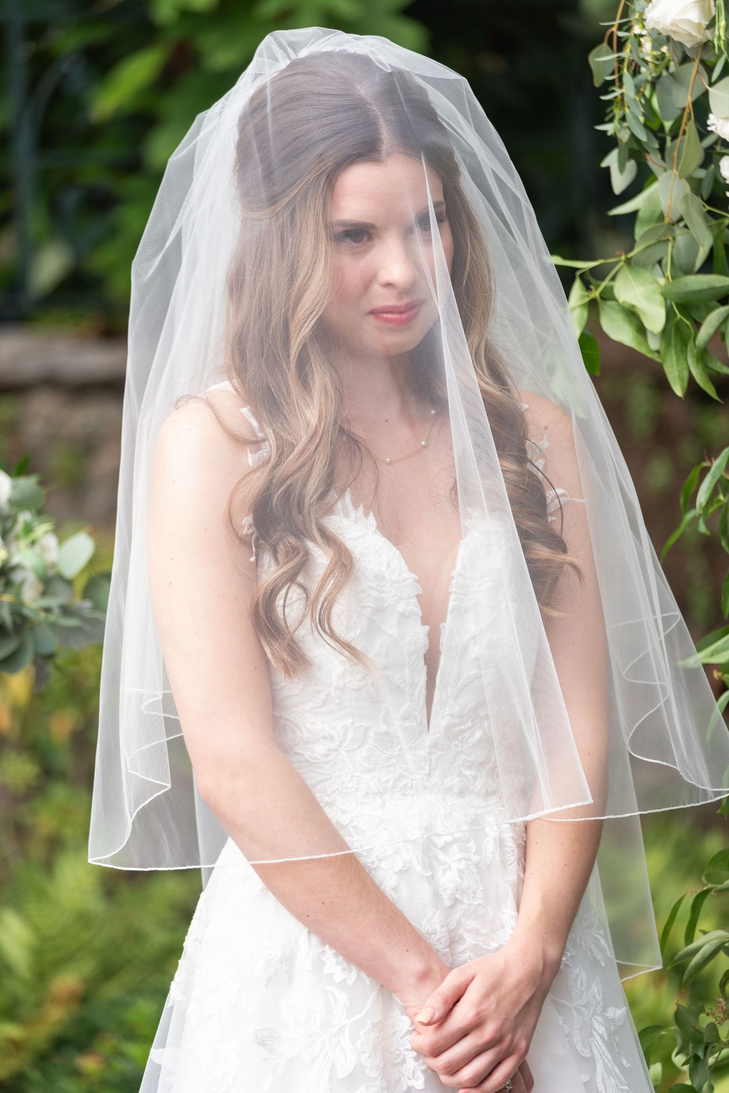 Beautiful bride in lace V-neck wedding gown and sheer veil poses outdoors surrounded by greenery.