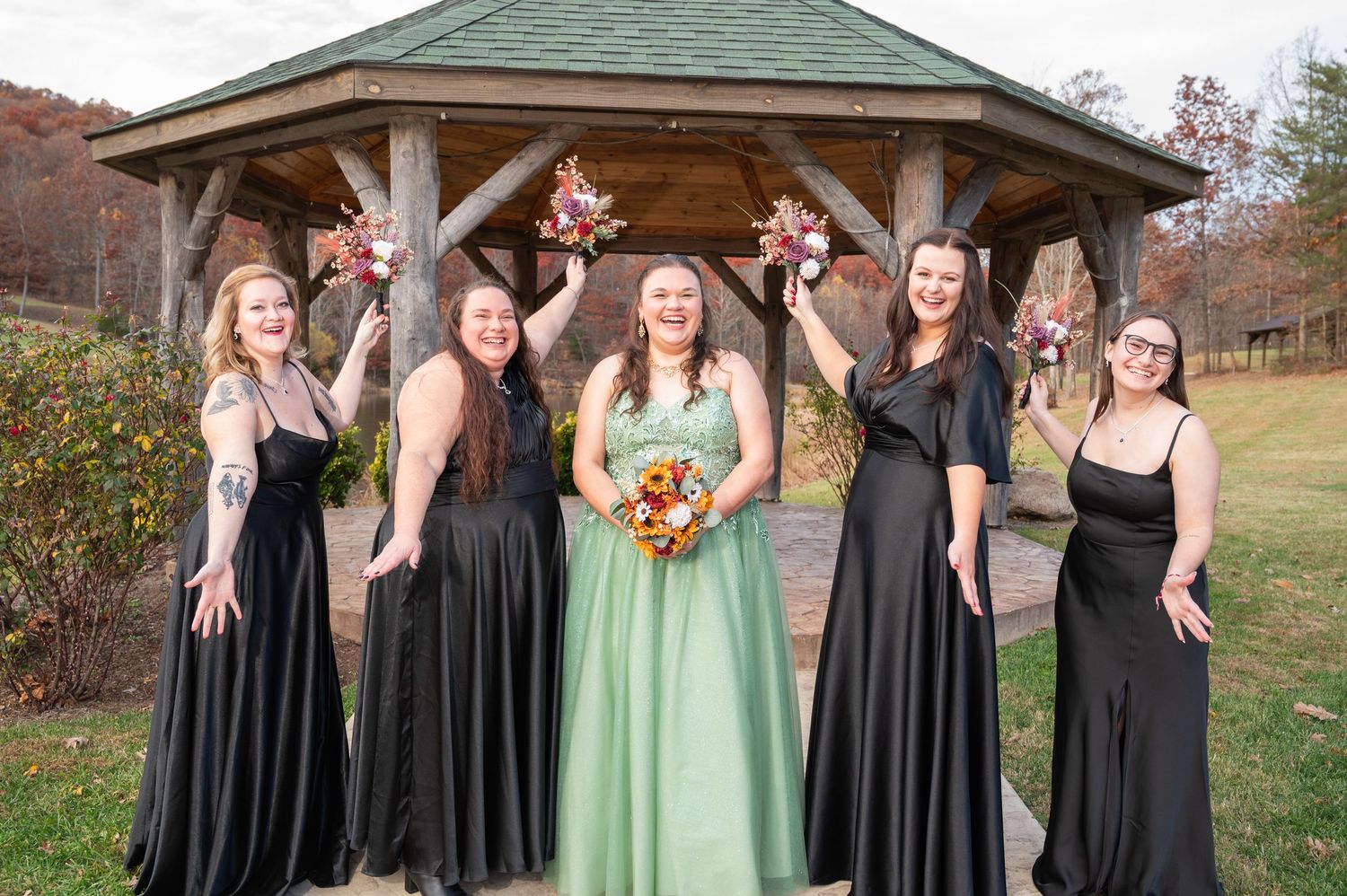 Bride in sage green gown with four bridesmaids in black dresses holding bouquets near rustic gazebo in autumn.
