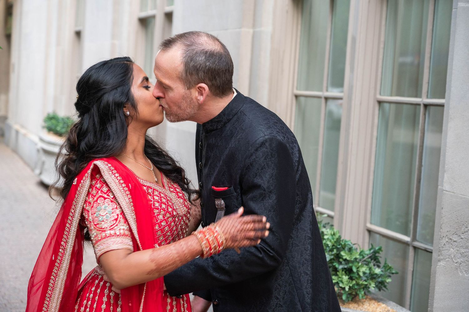 Couple sharing a romantic kiss, bride in red bridal lehenga with mehndi, groom in black sherwani, outdoor wedding photo.