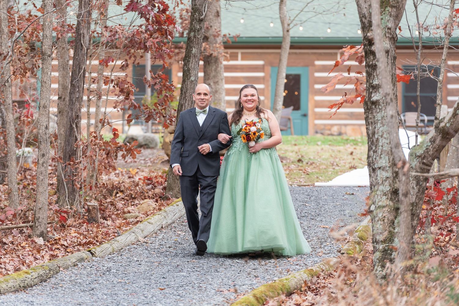 Bride in green gown walks with escort on gravel path past log cabin amid autumn foliage.