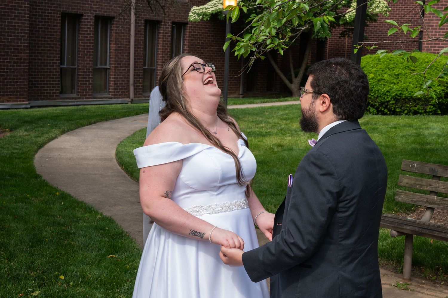 Joyful bride in off-shoulder white gown laughing while holding hands with groom outside brick church.