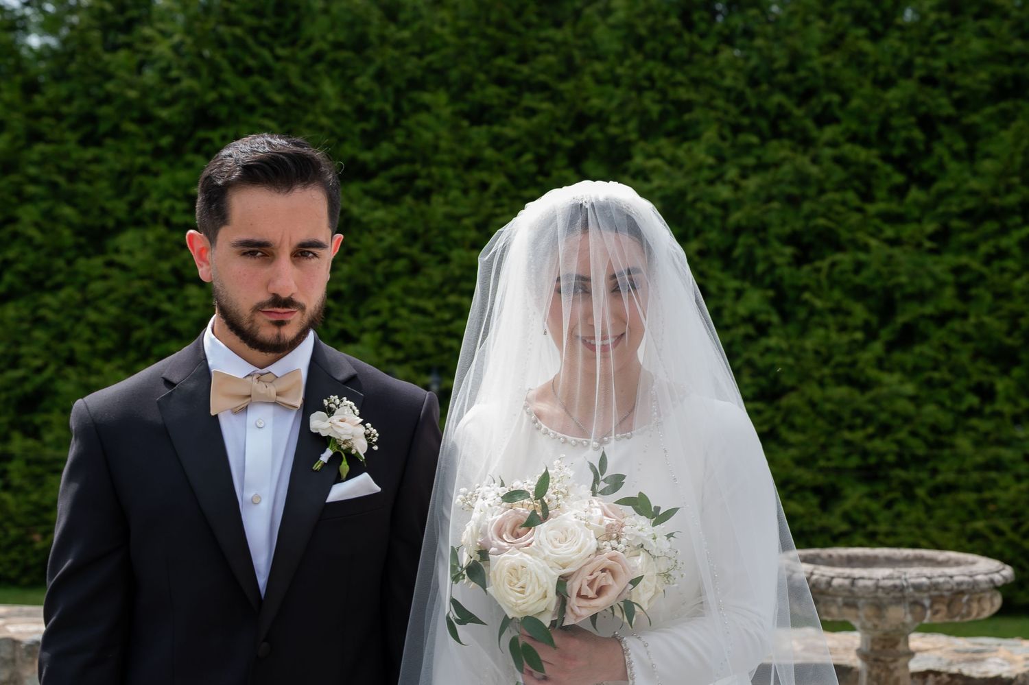 Bride and groom posing outdoors on wedding day, bride wearing veil holding white bouquet, groom in dark suit with bow tie.