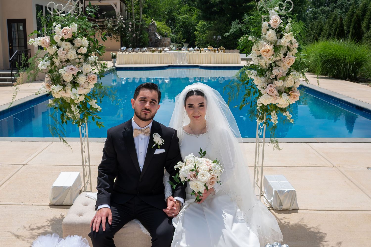 Bride and groom sit together in front of a floral arch and pool at an elegant outdoor wedding ceremony.