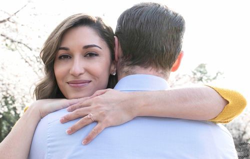 Smiling woman hugging her partner outdoors, showing off her diamond engagement ring.