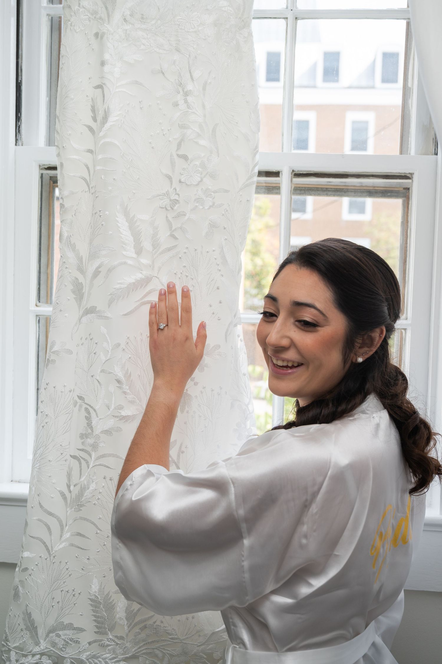 Bride in satin robe smiles while touching lace wedding dress hanging in window.