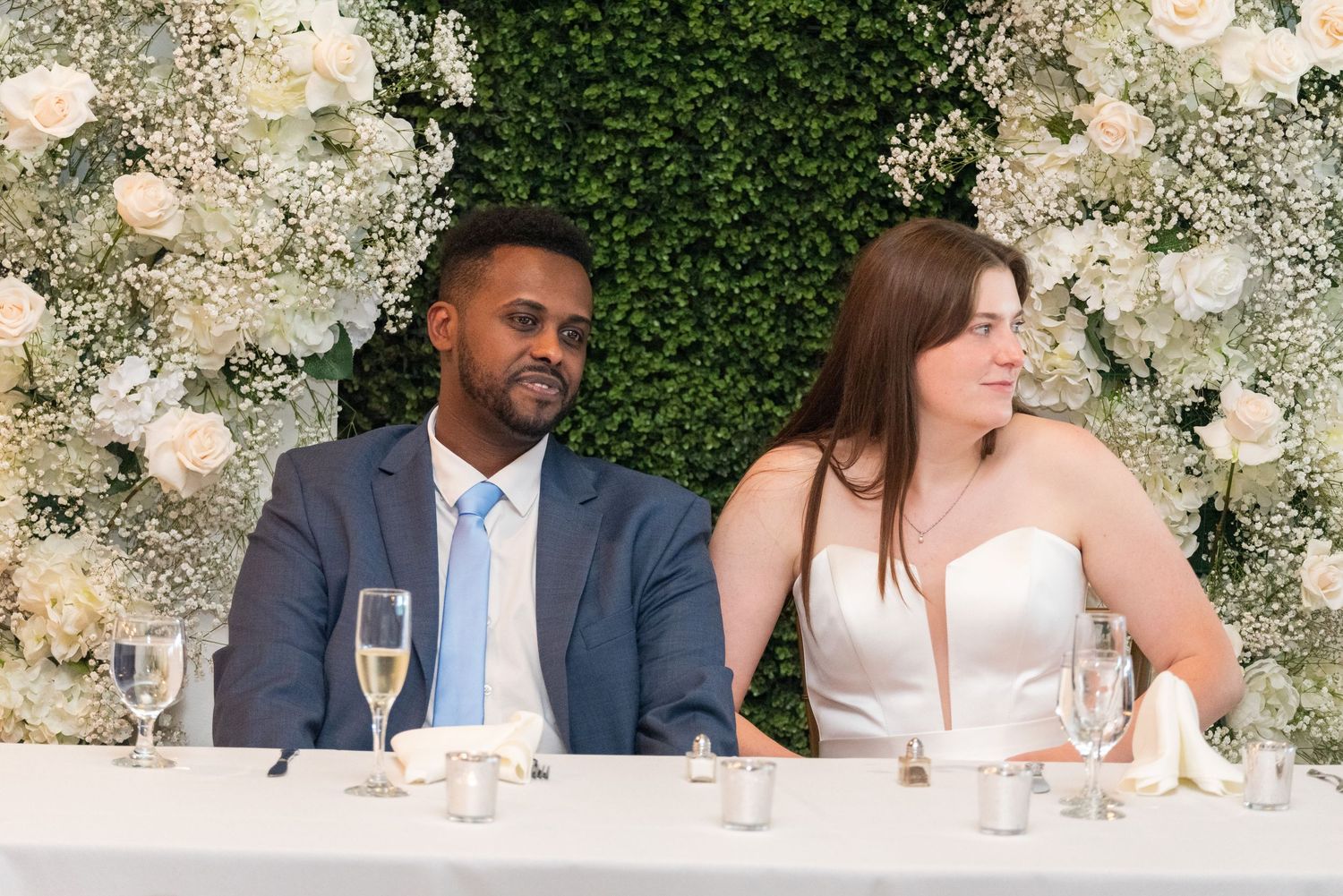 Newlywed couple sitting at wedding reception table with floral backdrop, groom in blue suit, bride in strapless gown.