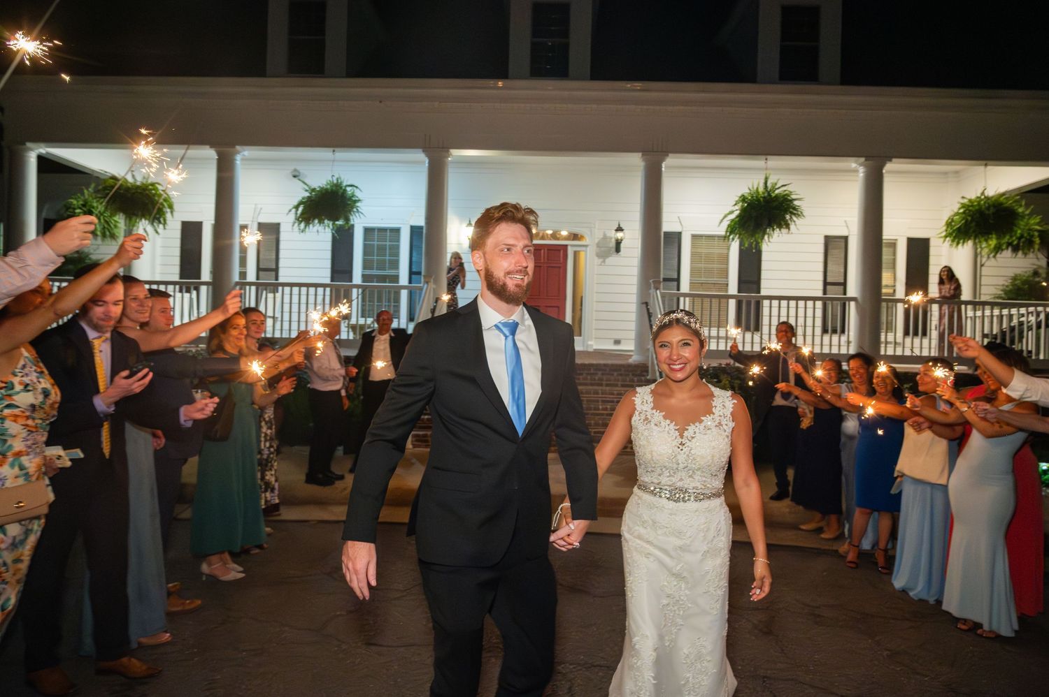 Bride and groom walk through sparkler exit at night wedding reception outside historic venue with guests cheering.