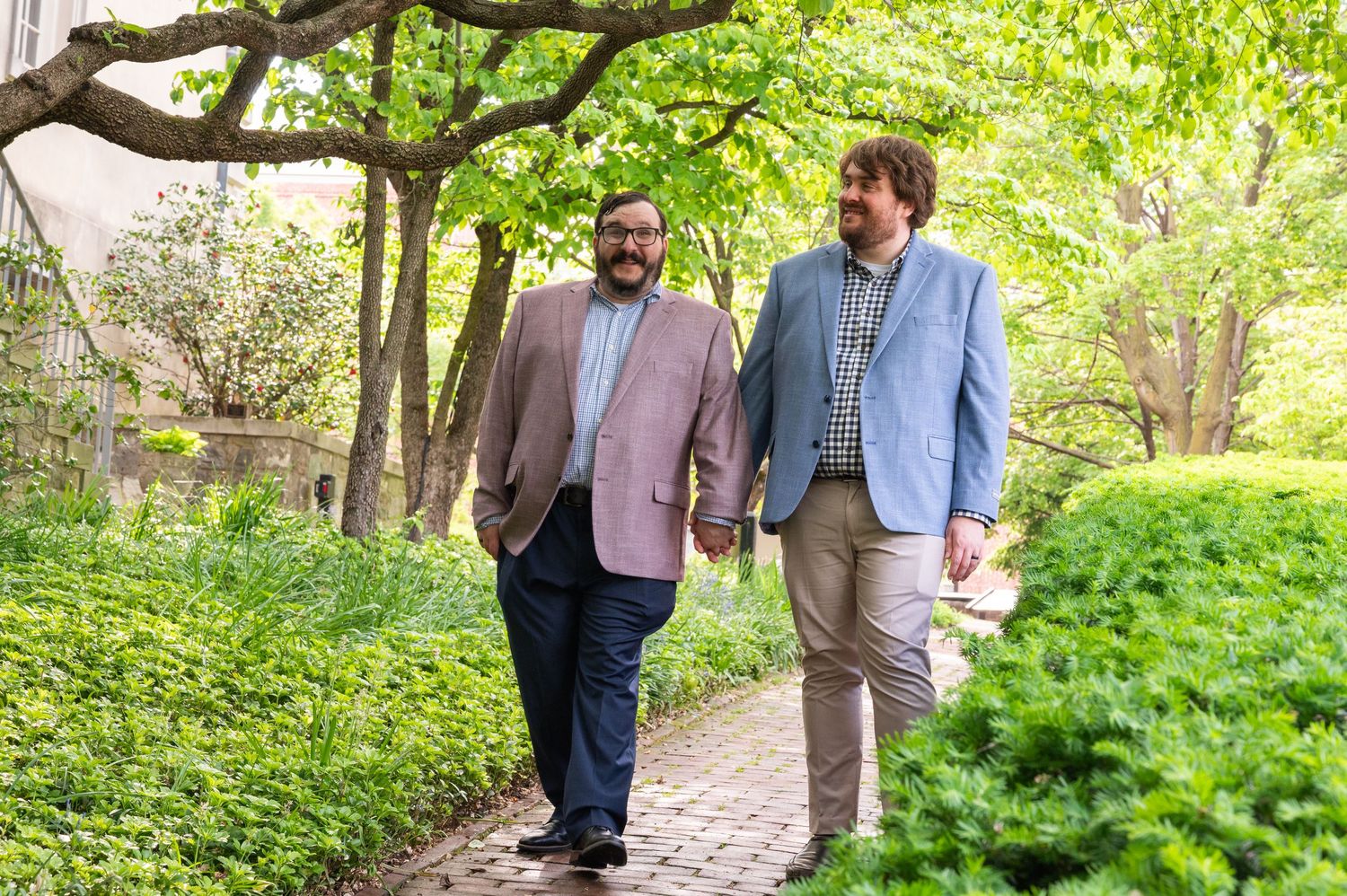 Two men holding hands walking along a brick garden path outdoors.