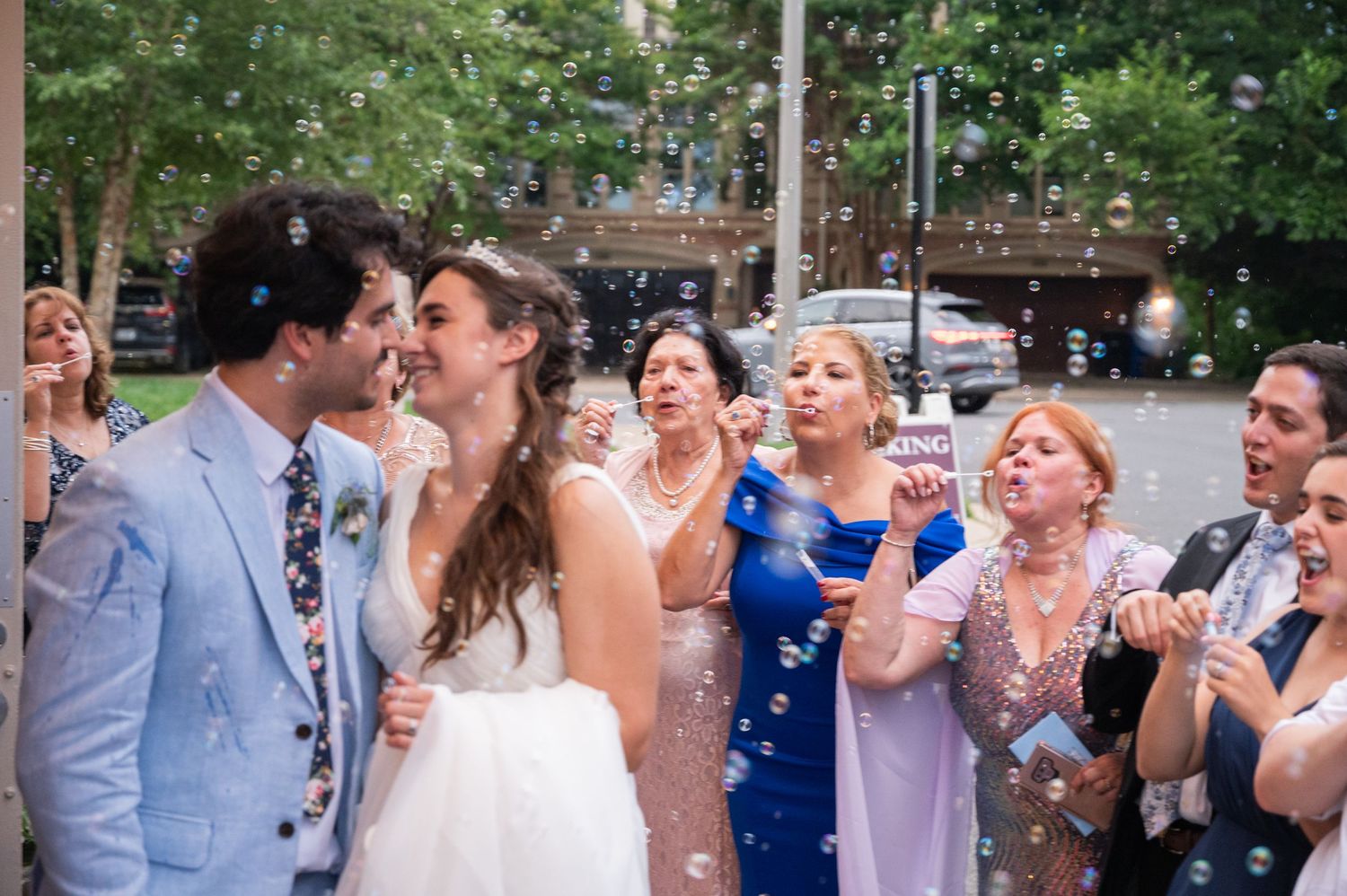 Bride and groom kissing amid bubbles surrounded by celebrating guests on city street.