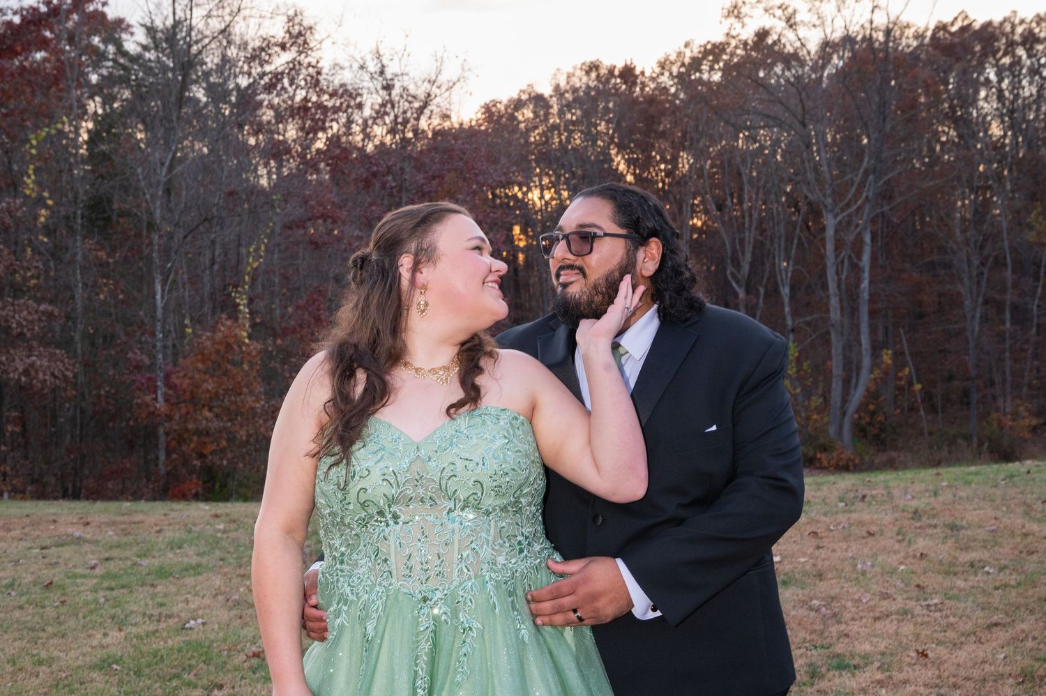 Couple in formal wear, woman in green gown and man in black suit, smiling outdoors in autumn setting.