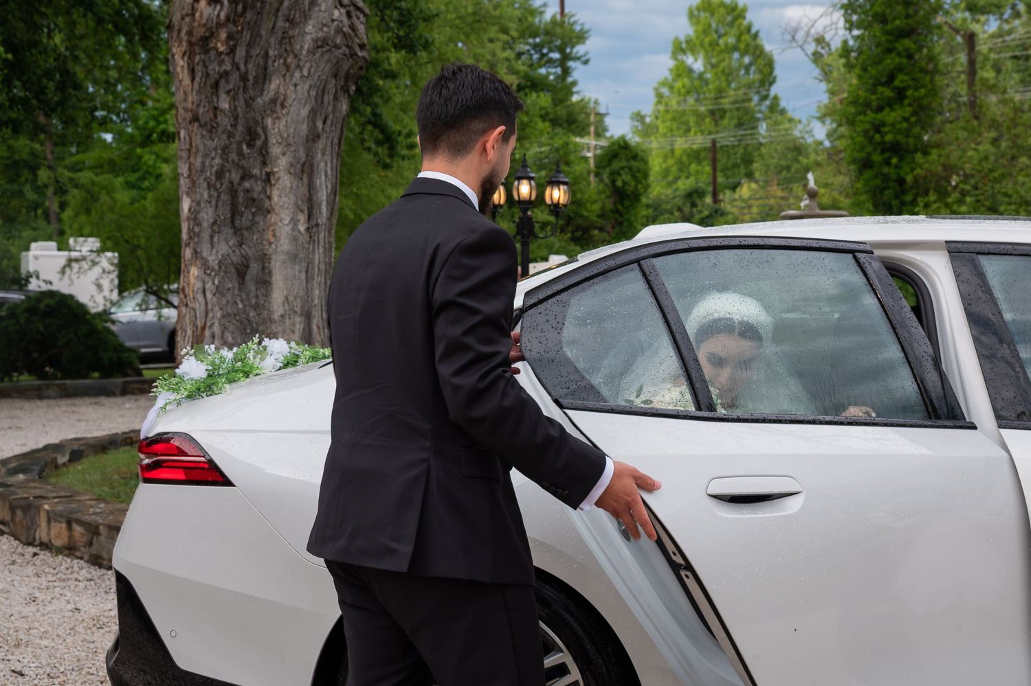 Groom in black suit opening white wedding car door for bride, elegant outdoor ceremony setting.