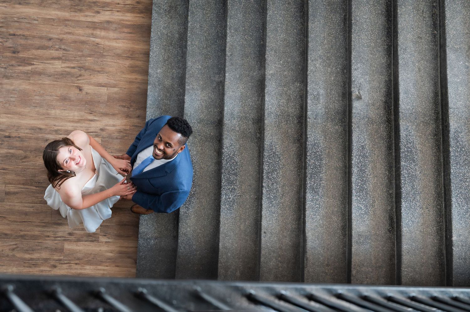 Aerial view of smiling interracial couple holding hands on concrete stairs, looking up at camera.